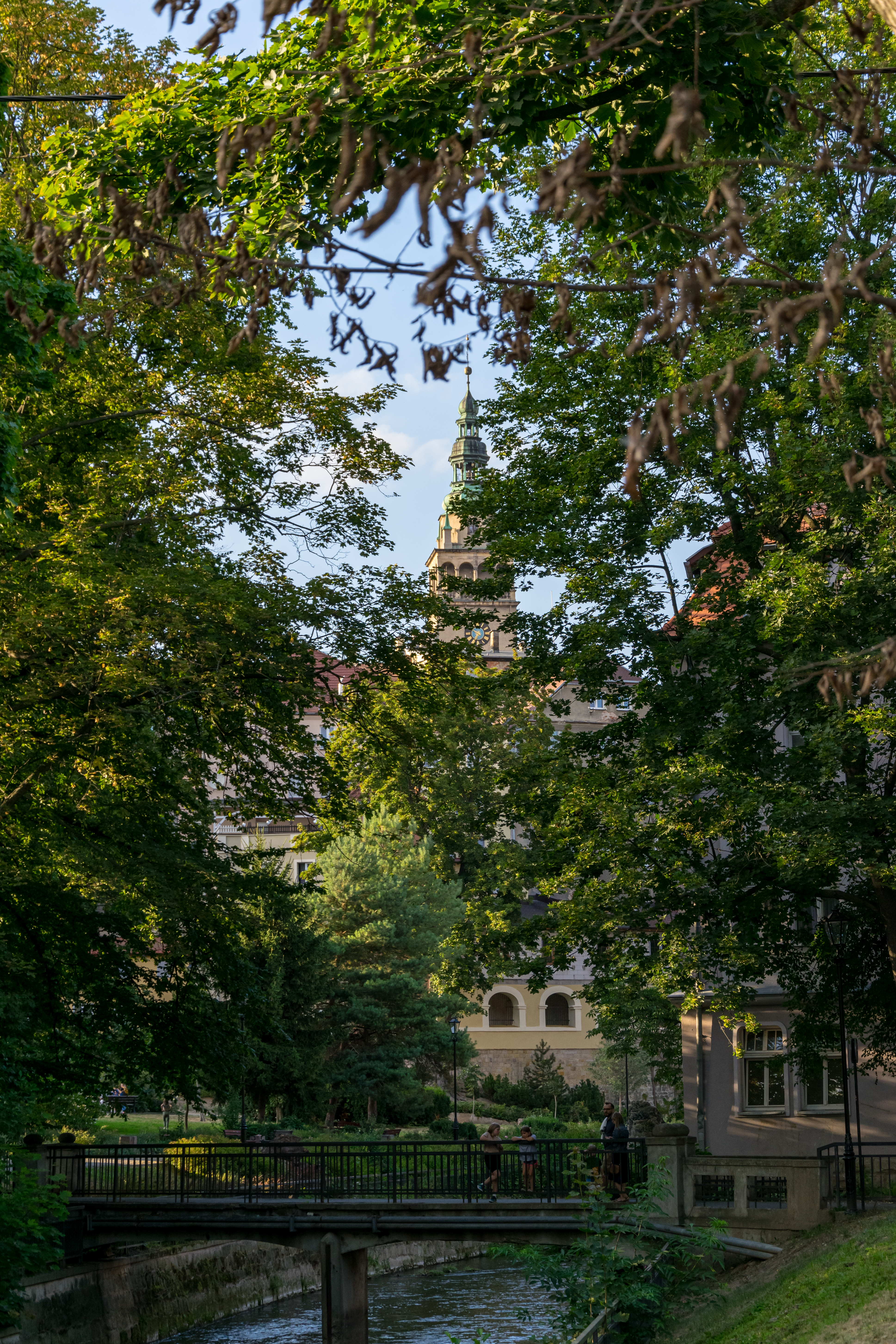 small river in park, with a bunch of people on a bridge, and a church hidden behind trees in the background