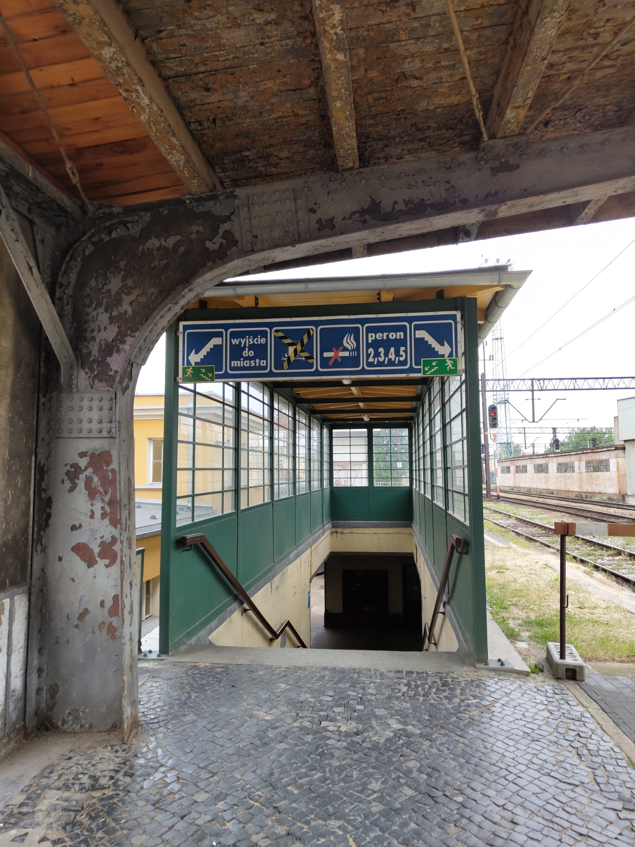 Photo of platform signage showing an arrow with stairs to the left, "exit to city", something crossed out by tape, a cigarette crossed out by a red cross, "platform 2,3,4,5" and another arrow with stairs, but to the right