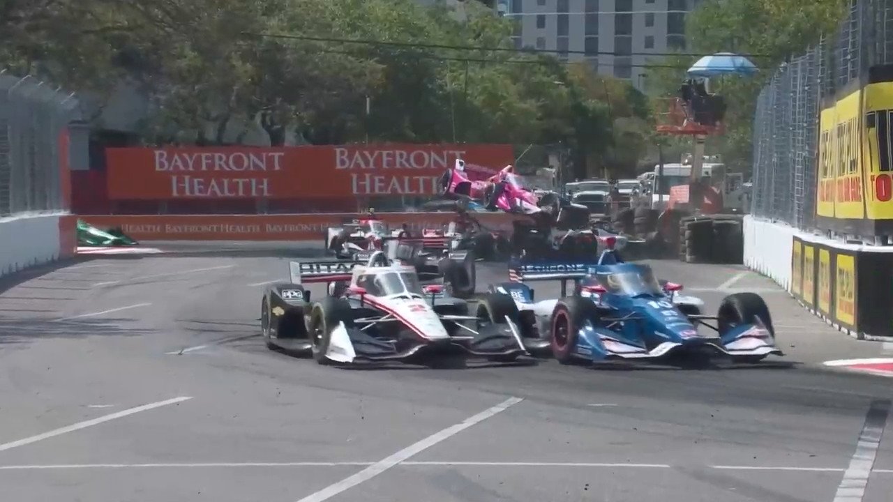 screenshot of an indycar race, pink car in the background flying in the air