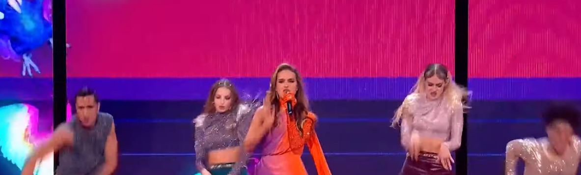 polish eurovision singer and 2 dancers with a background that looks a bit like a bi flag