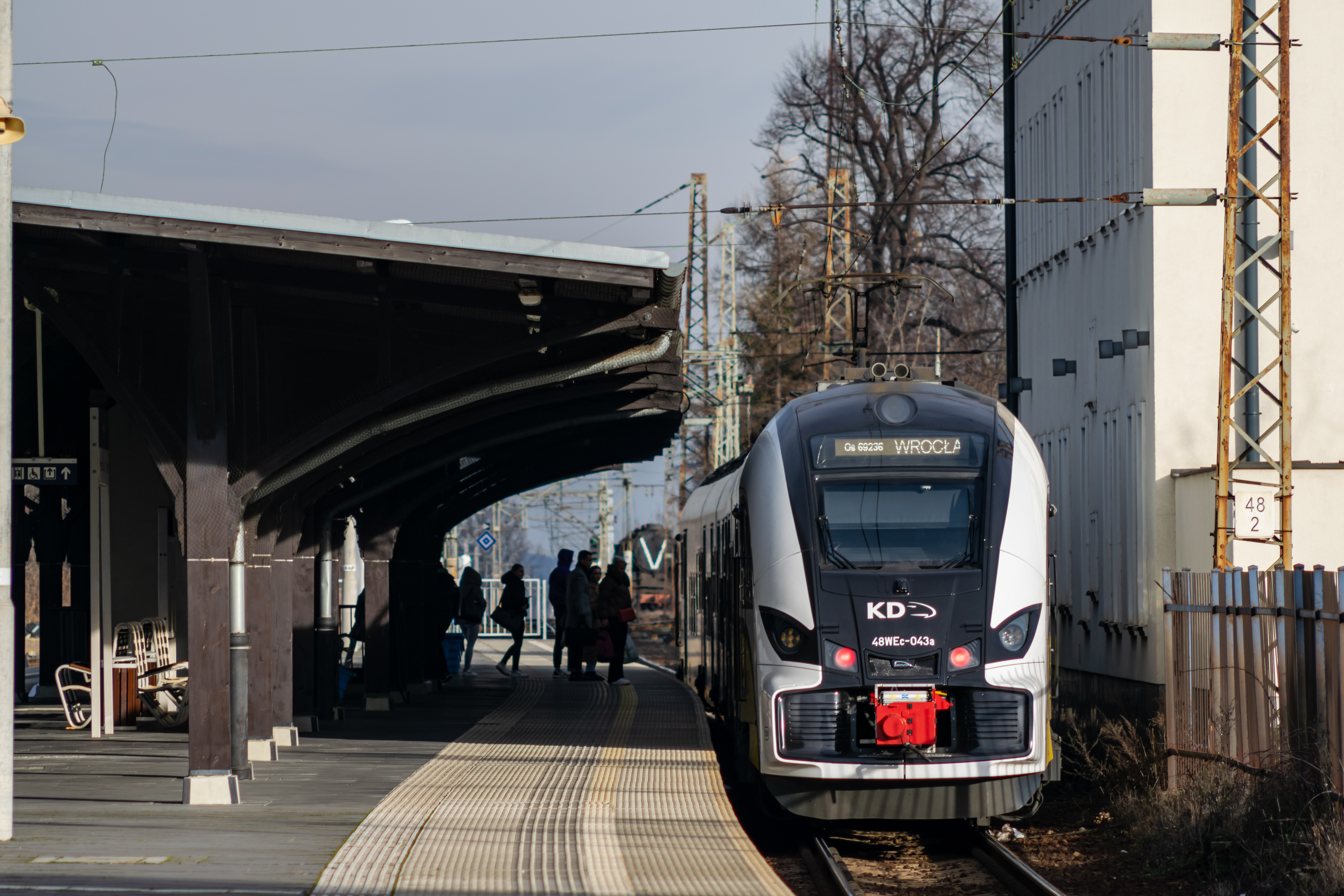 yellow-black-white train standing at a sheltered platform with people waiting to get on board