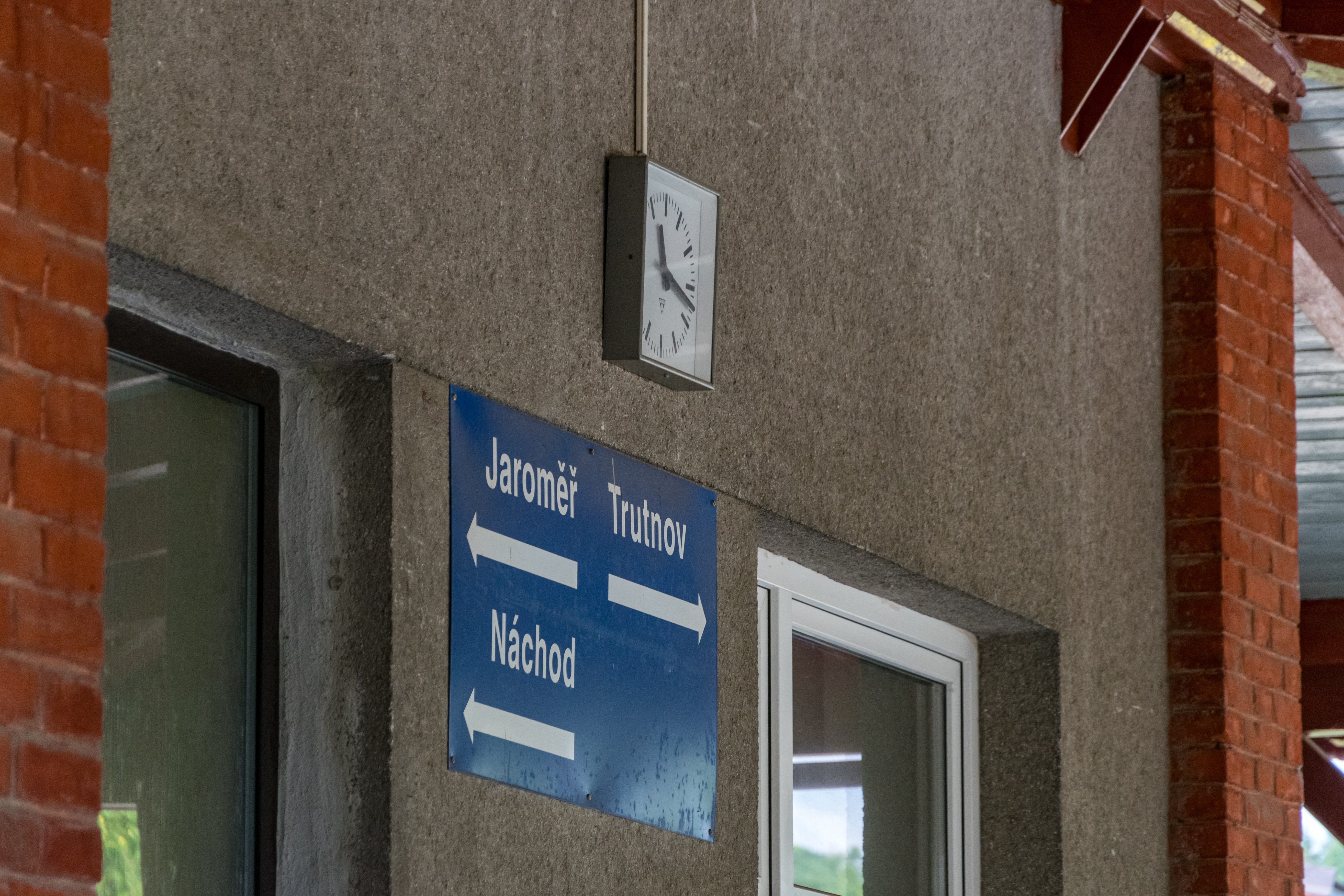 grey concrete wall of train station in Starkoč with square clock and a directional sign with arrows pointing towards Jaroměř, Trutnov and Náchod. Jaroměř and Náchod are to the left, while Trutnov is to the right. Wall is bordered by red brick columns