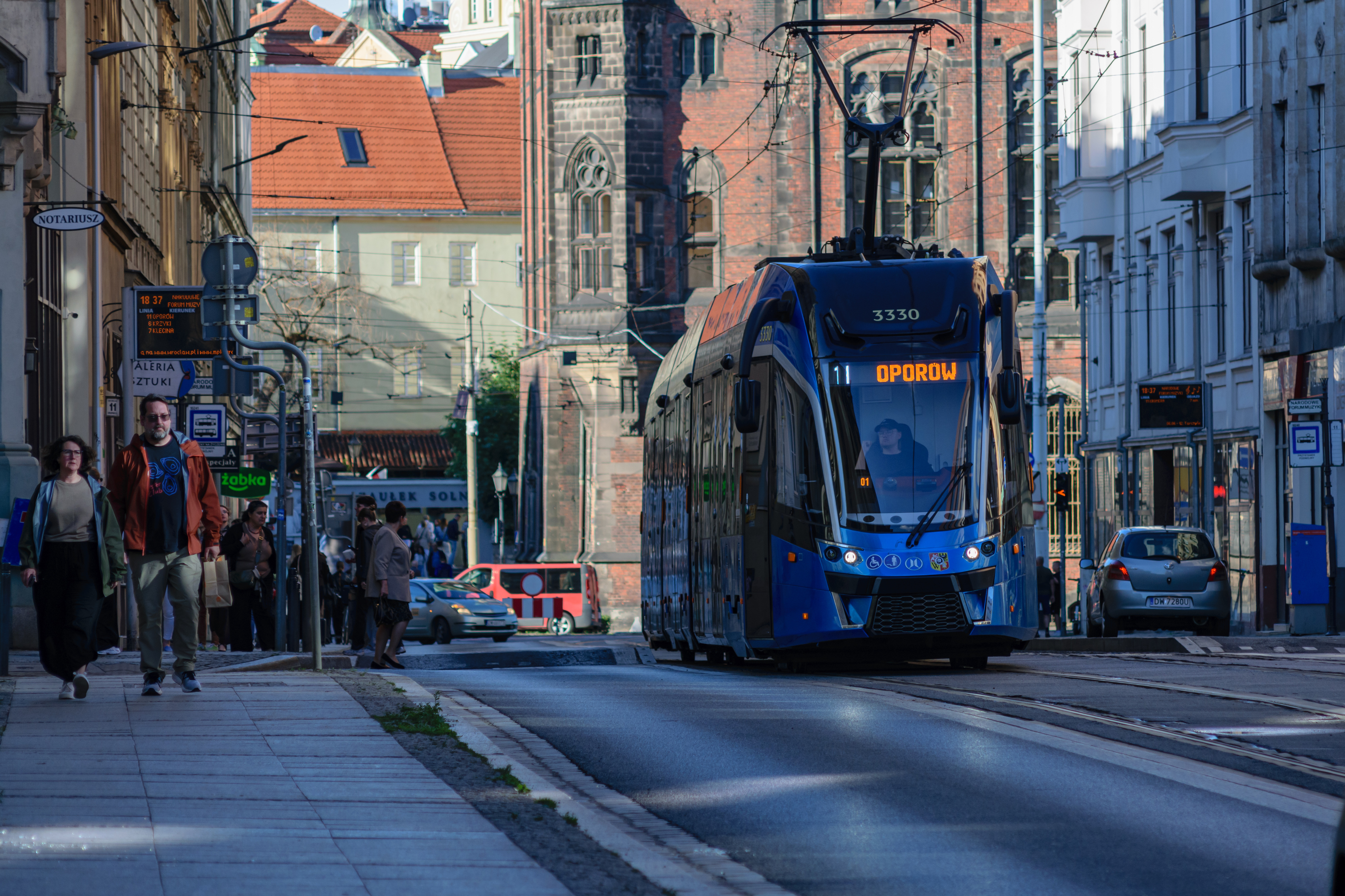 Blue tram driving of a tram stop in old town, there are people walking on the sidewalk