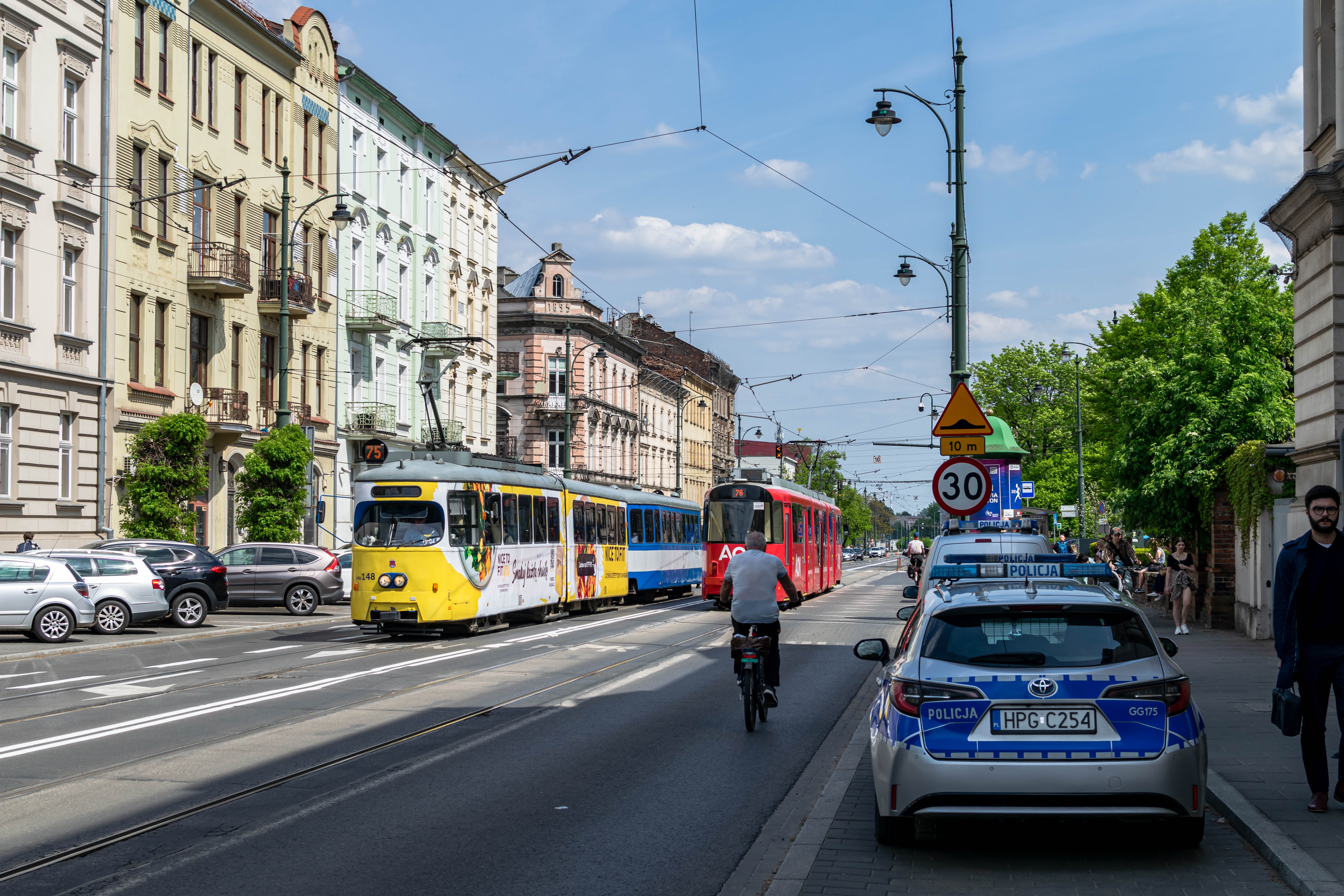 Old tram from Vienna with ads, pulling another coach, and some other modernised Düwag/SGP tram going in opposite direction, obstructed by a cyclist. There’s a lot of old buildings from around 19th century