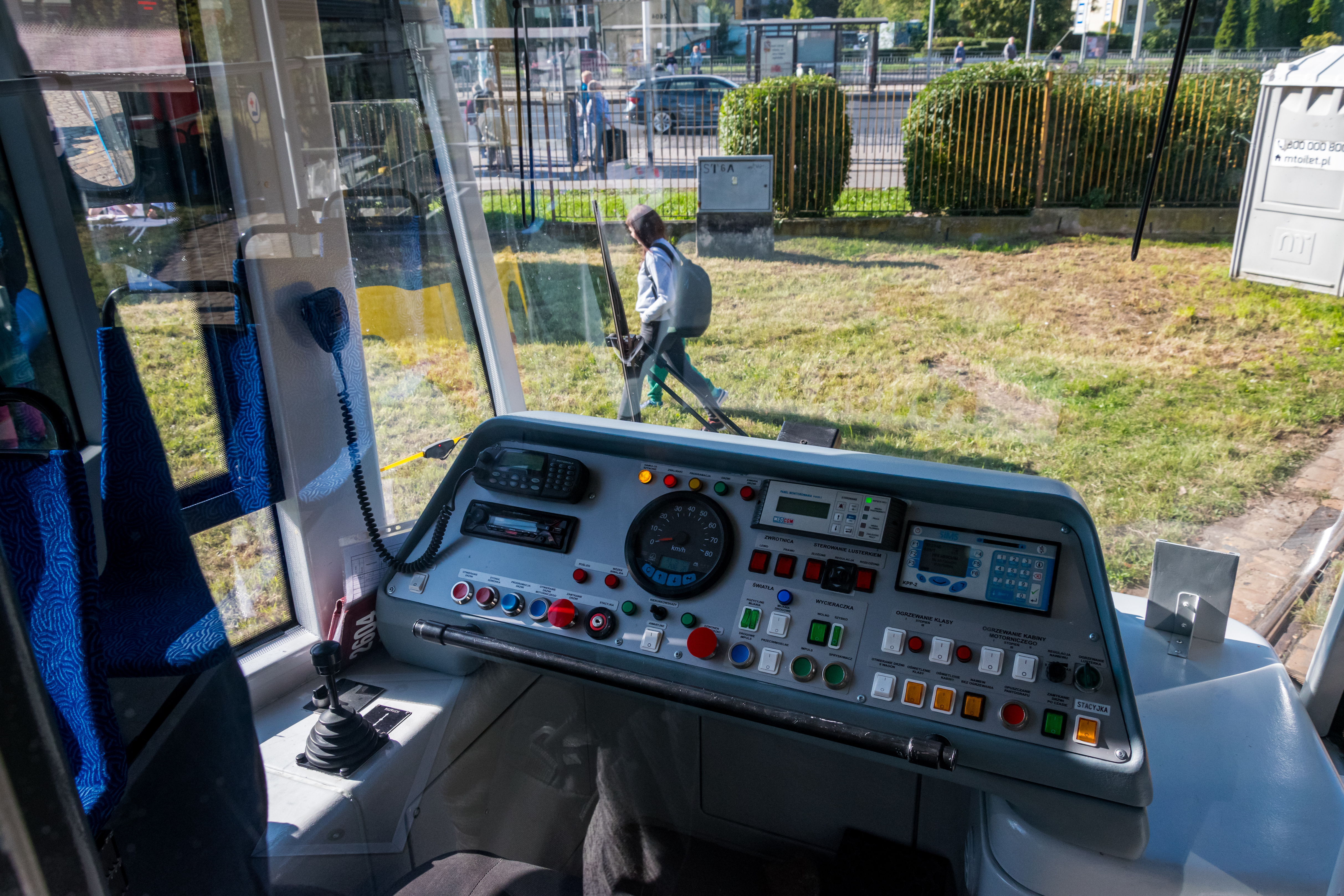 a tram cockpit with various controls, view through the front window shows a person walking outside on grass
