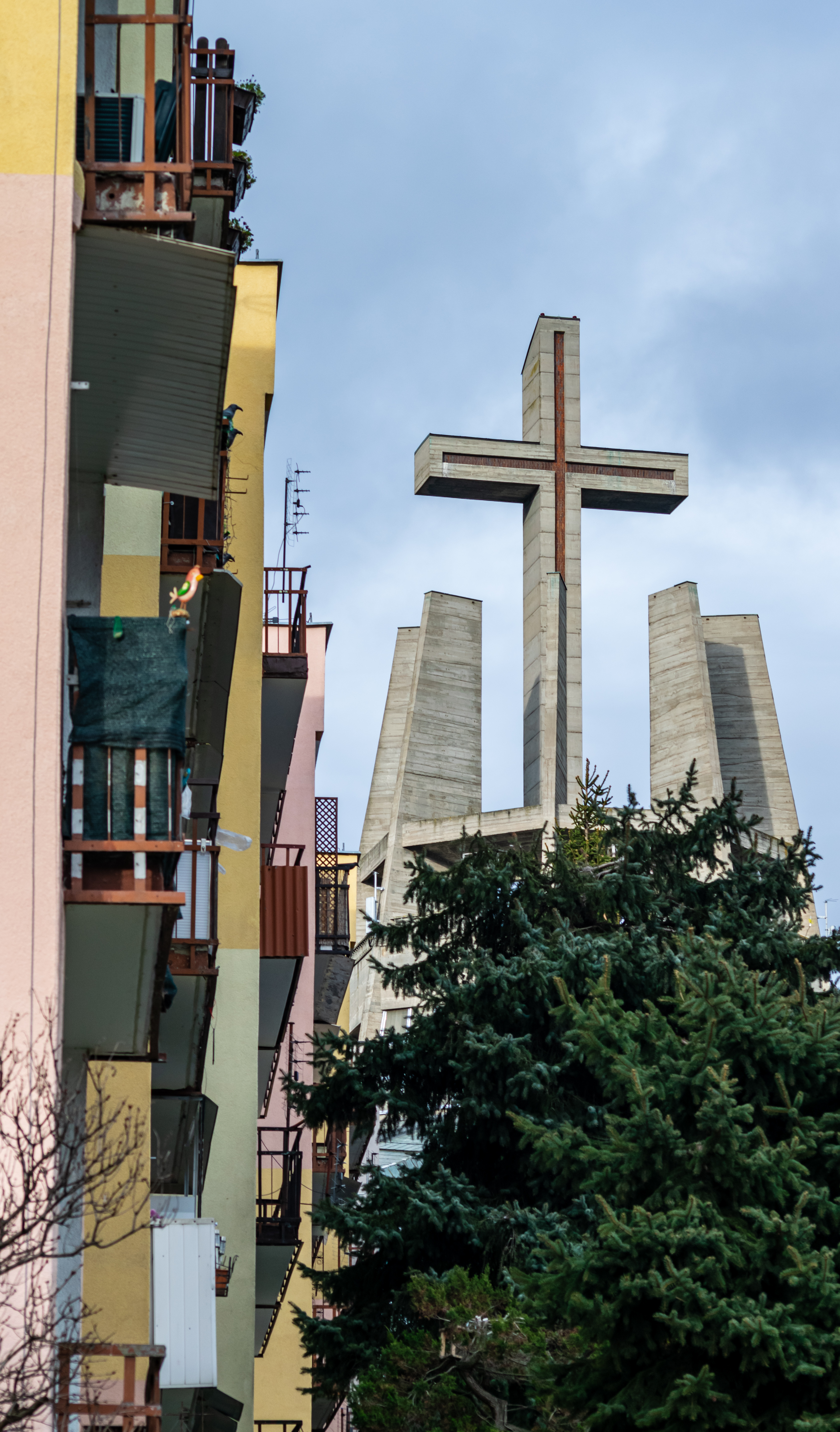 big cross hiding behind a tree, next to a commie block viewed from side