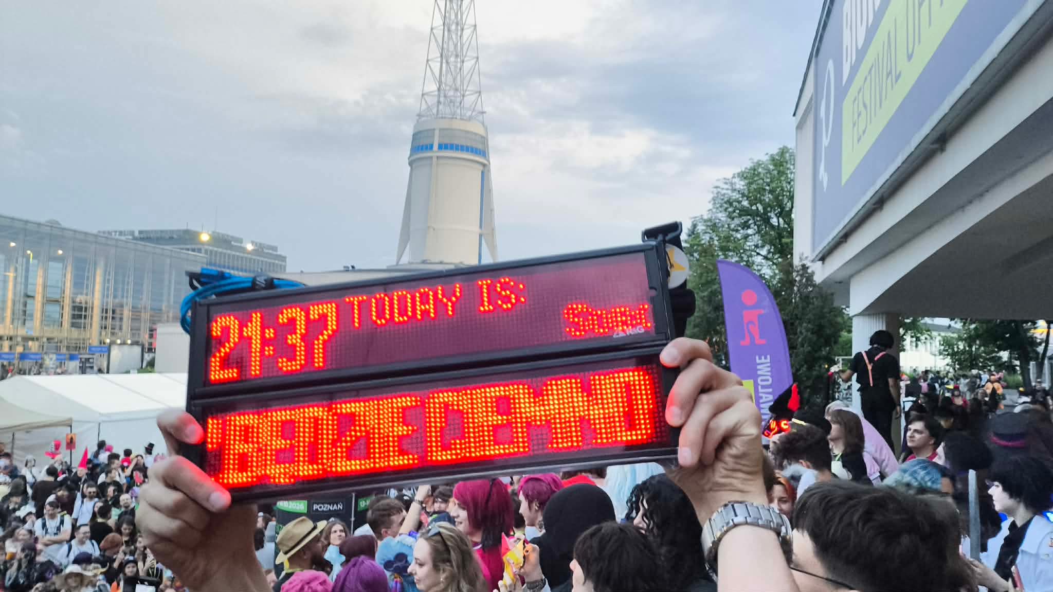 Two R&G train displays being held in the air displaying the hour 21:37 and a scrolling text "zaraz będzie ciemno"