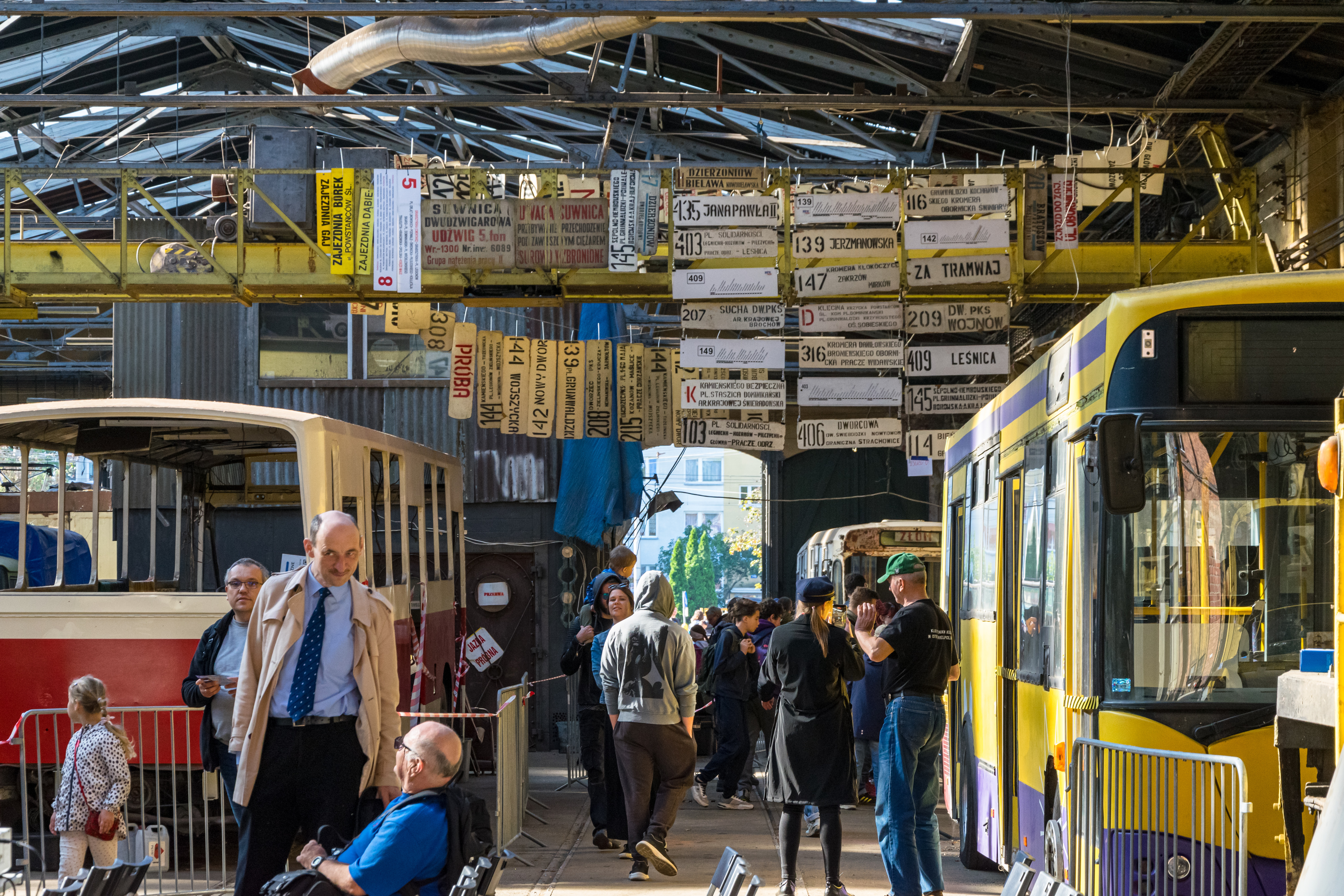 people walking around a tram depot with buses and a lot of old destination signs hanging from the ceiling