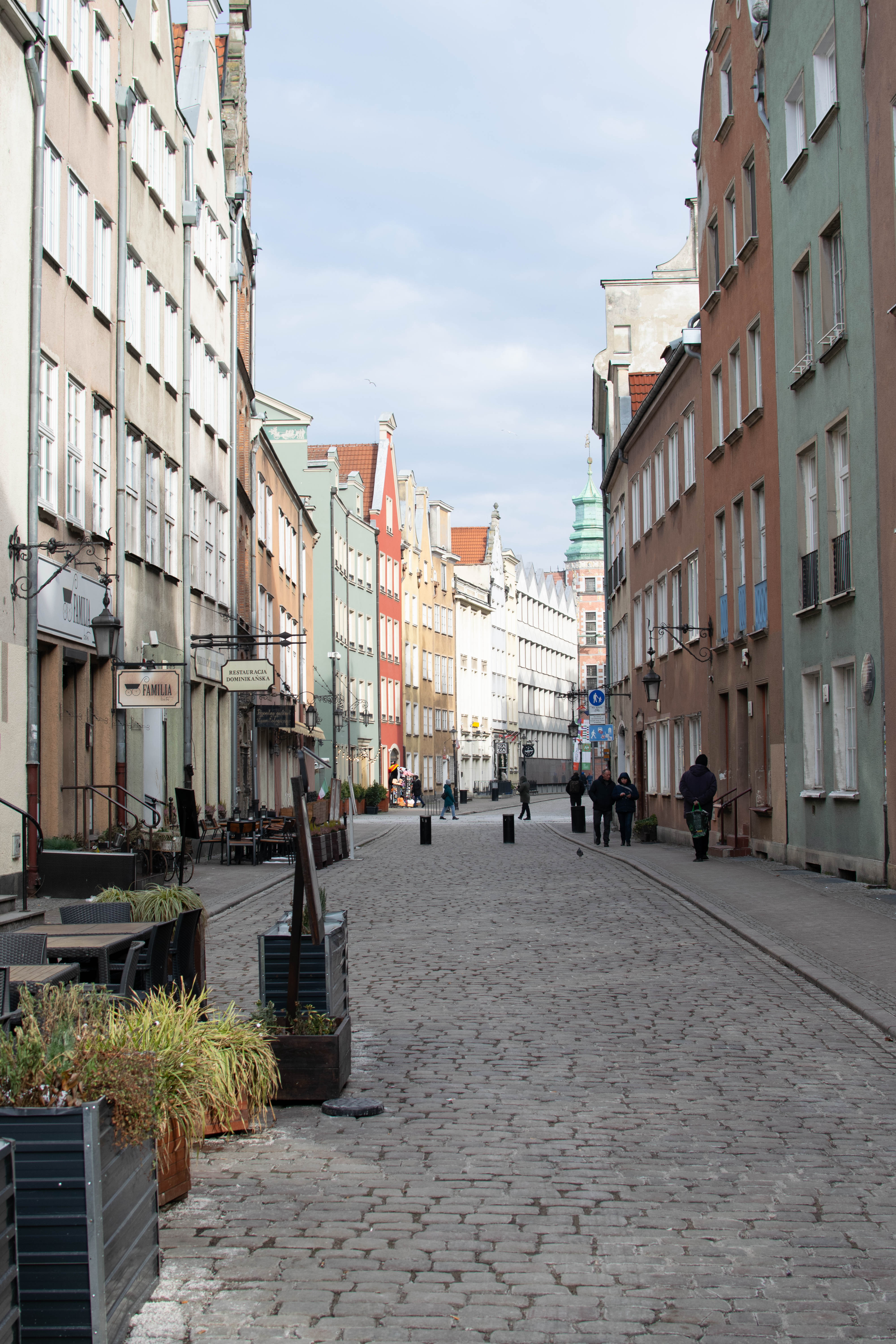 cobblestone street with colorful townhouses on both sides, there's a bunch of cafe gardens on the left and few people walking 