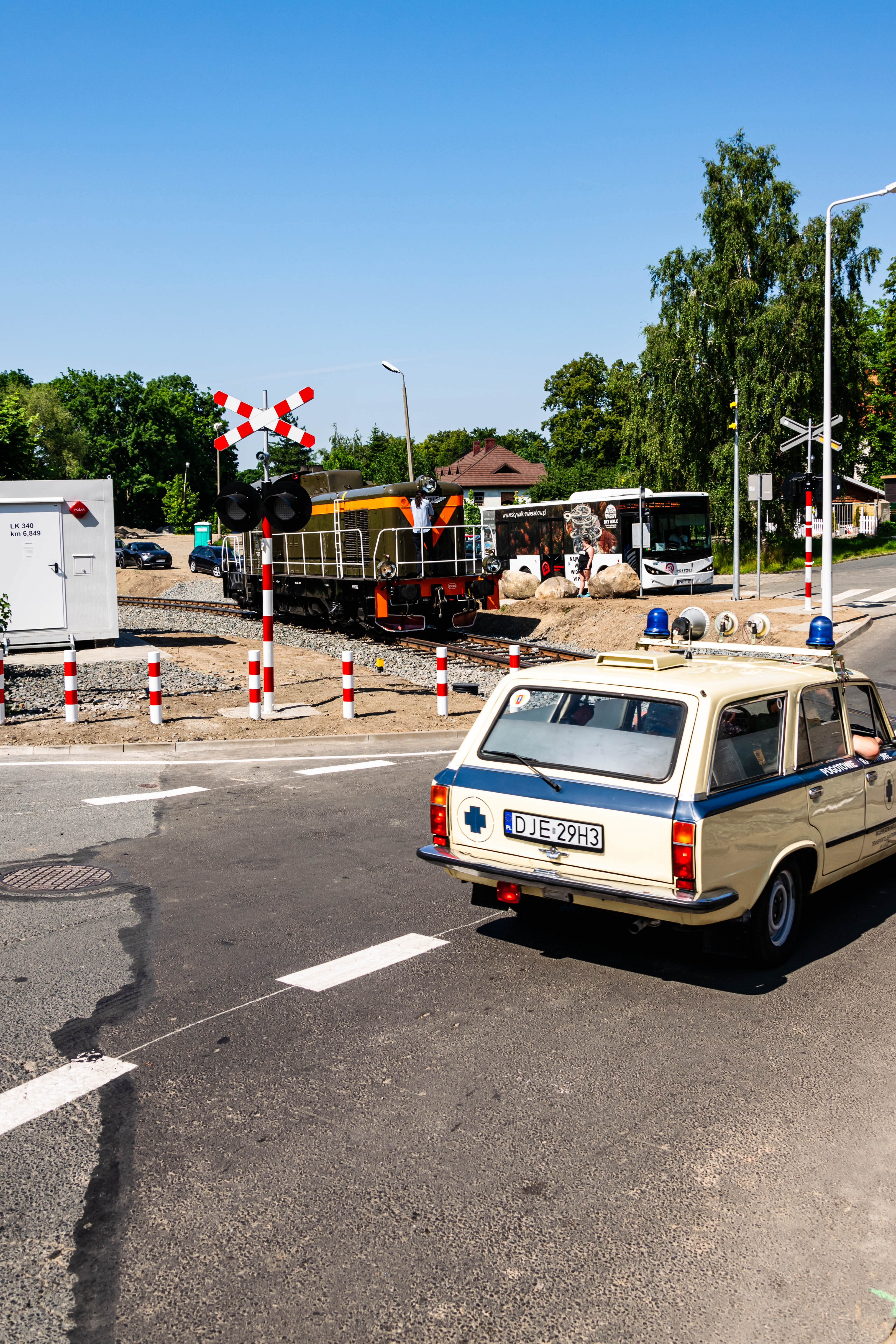 restored fiat 125p with medical emergencies livery stopped at a railway crossing, with a restored SP42 diesel locomotive on it, shunting, there's a modern isuzu bus stopped behind the locomotive