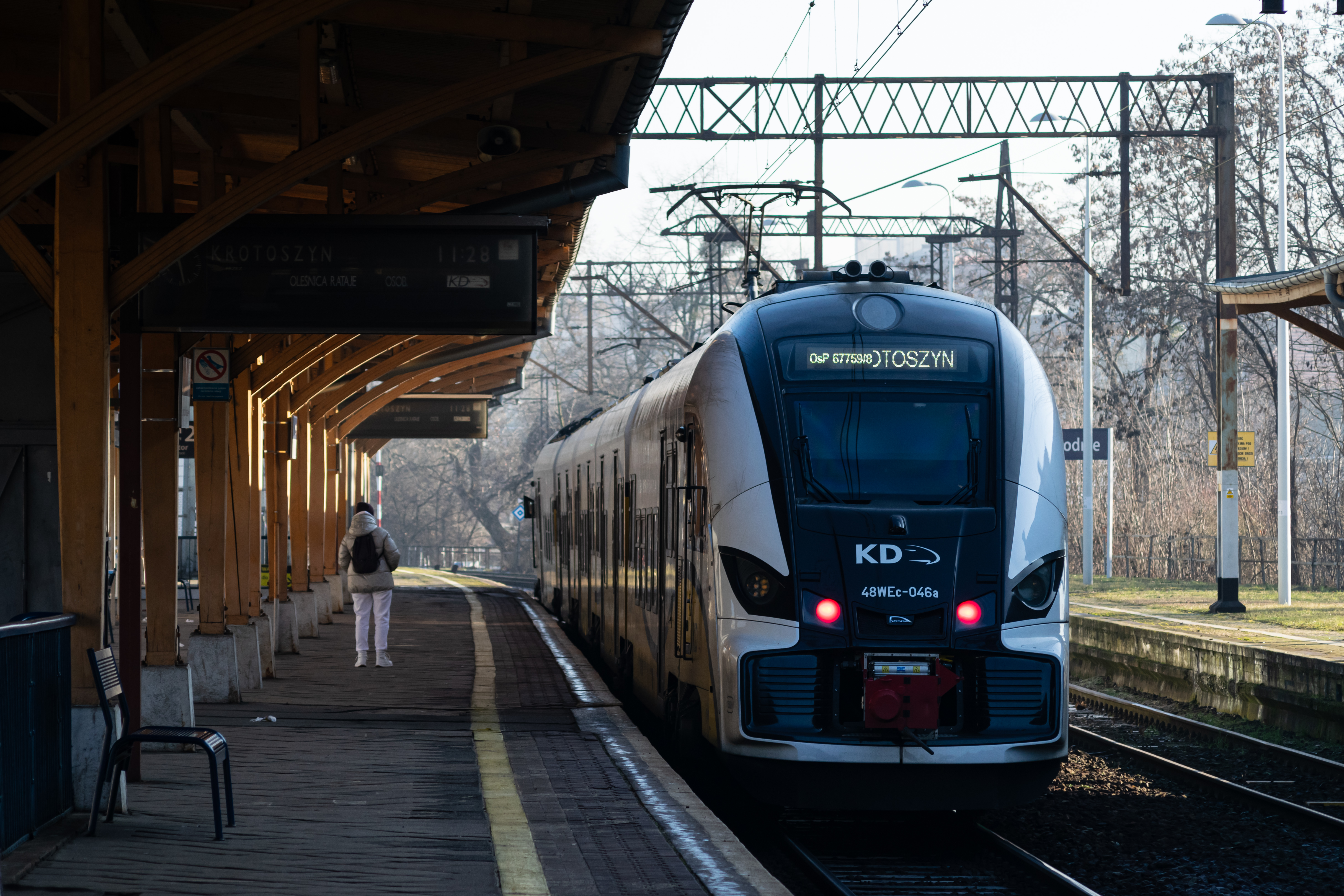yellow-white train standing next to a platform with wooden shelter on whole length that features flip displays, there's a singular person standing,