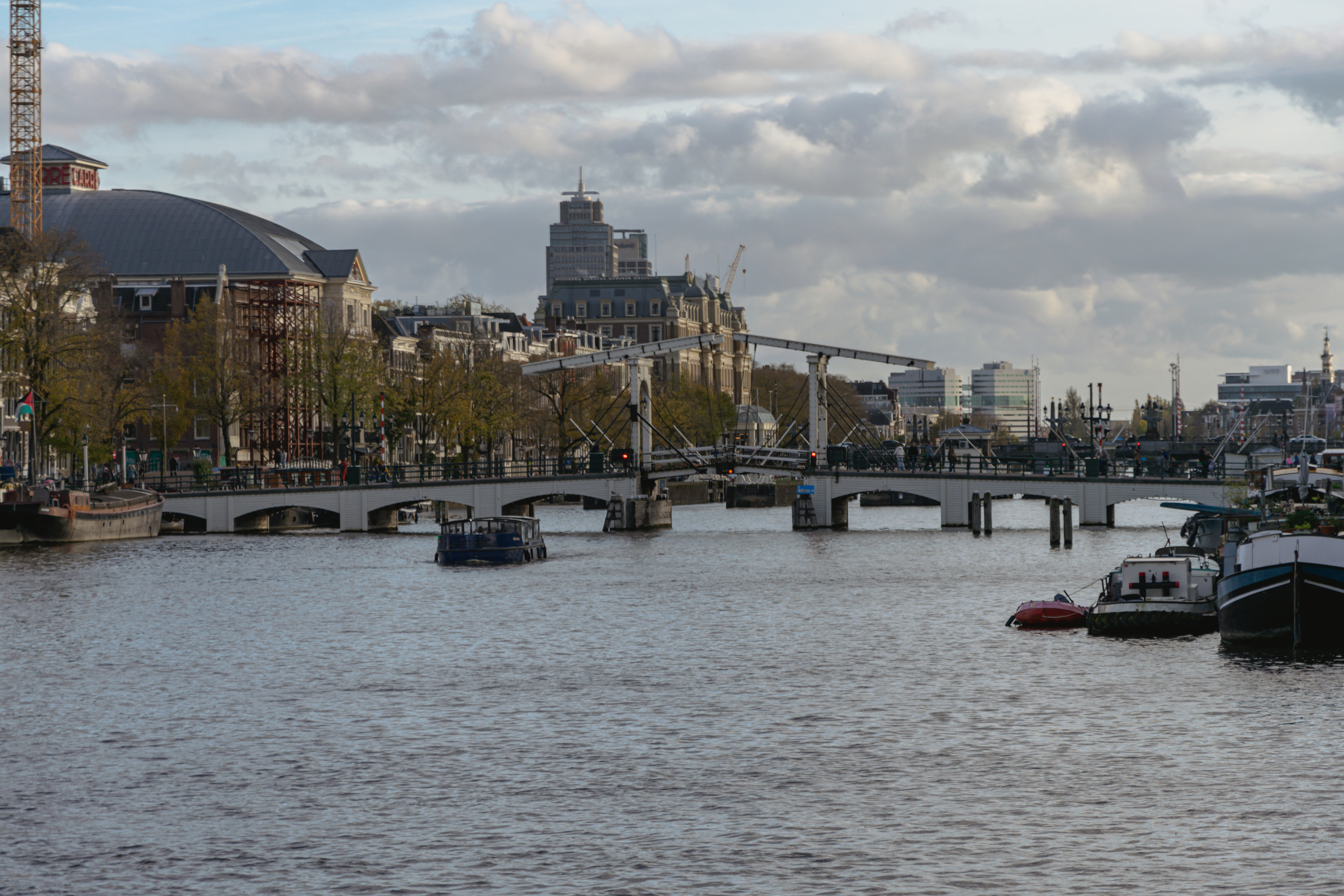 A view of a river with boats, a bridge with a drawbridge mechanism, and city buildings in the background under partly cloudy skies