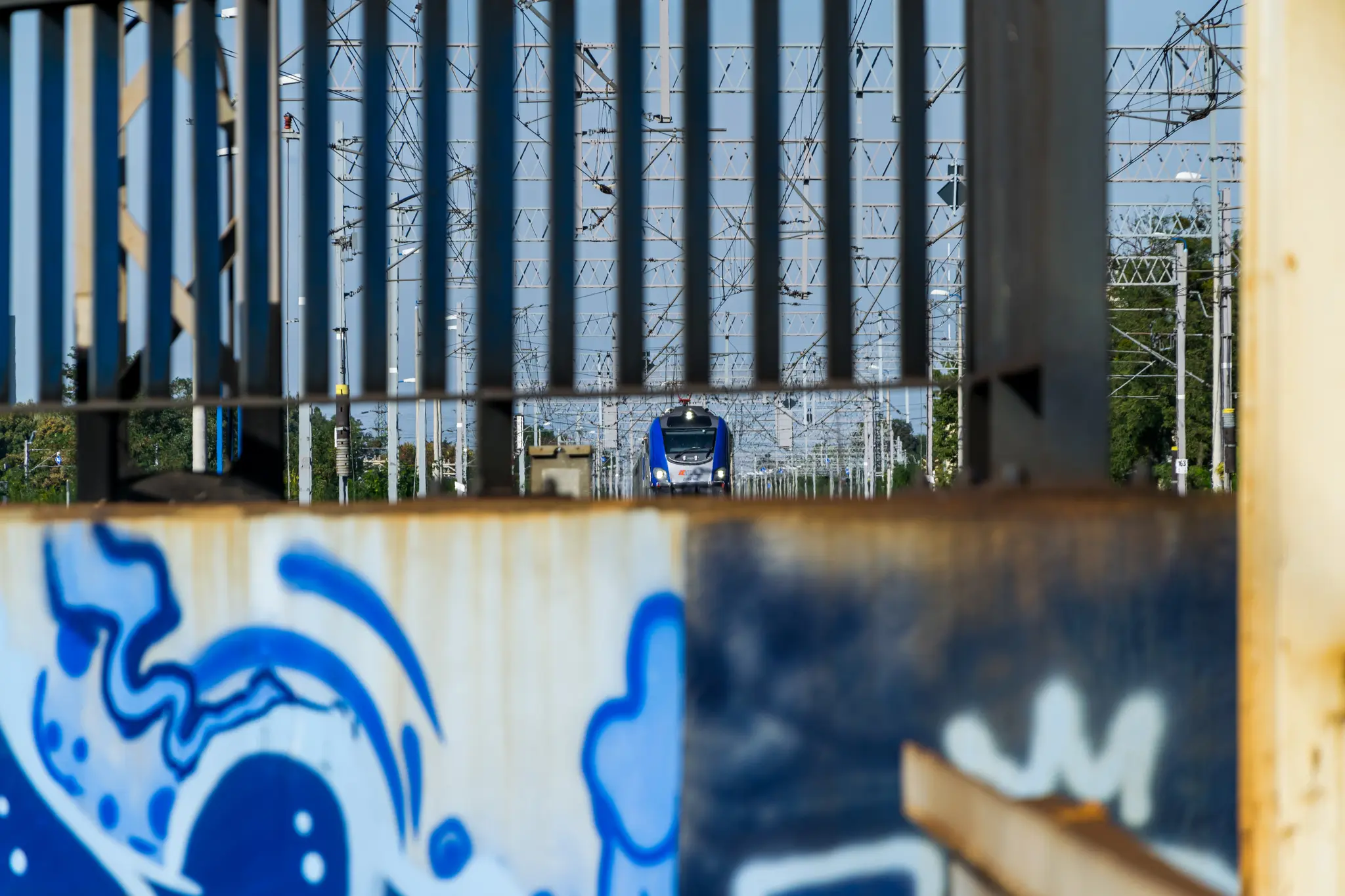 Blue-silver Griffin EU160 locomotive with a bunch of coaches arriving at the station, viewed through a fence