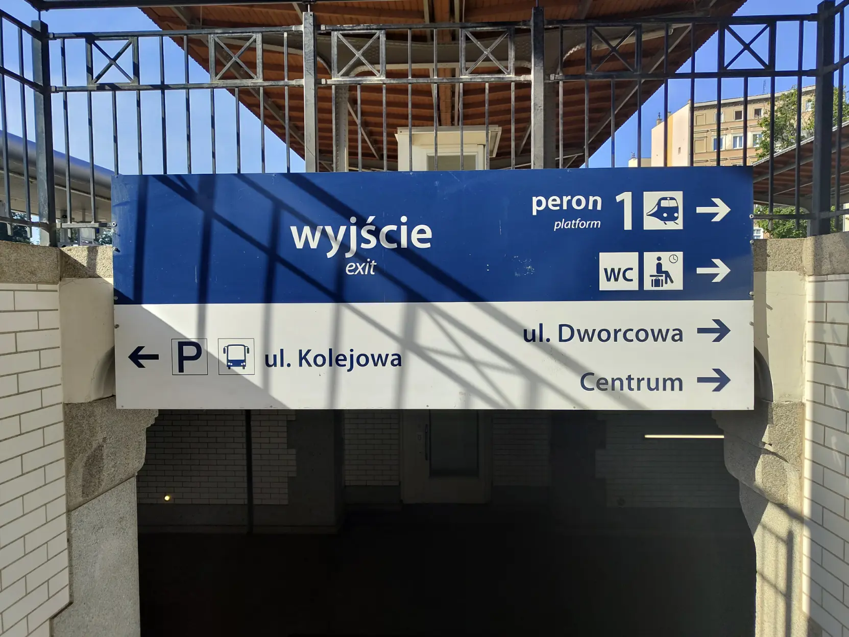 two-toned sign, dark blue background with white lettering on upper half, and white background with dark blue lettering on lower half. Upper half informs about exit from the station, way to the platform 1, alongside way to the wc and waiting room. Lower half informs about ways to city centre, and a coach bus station