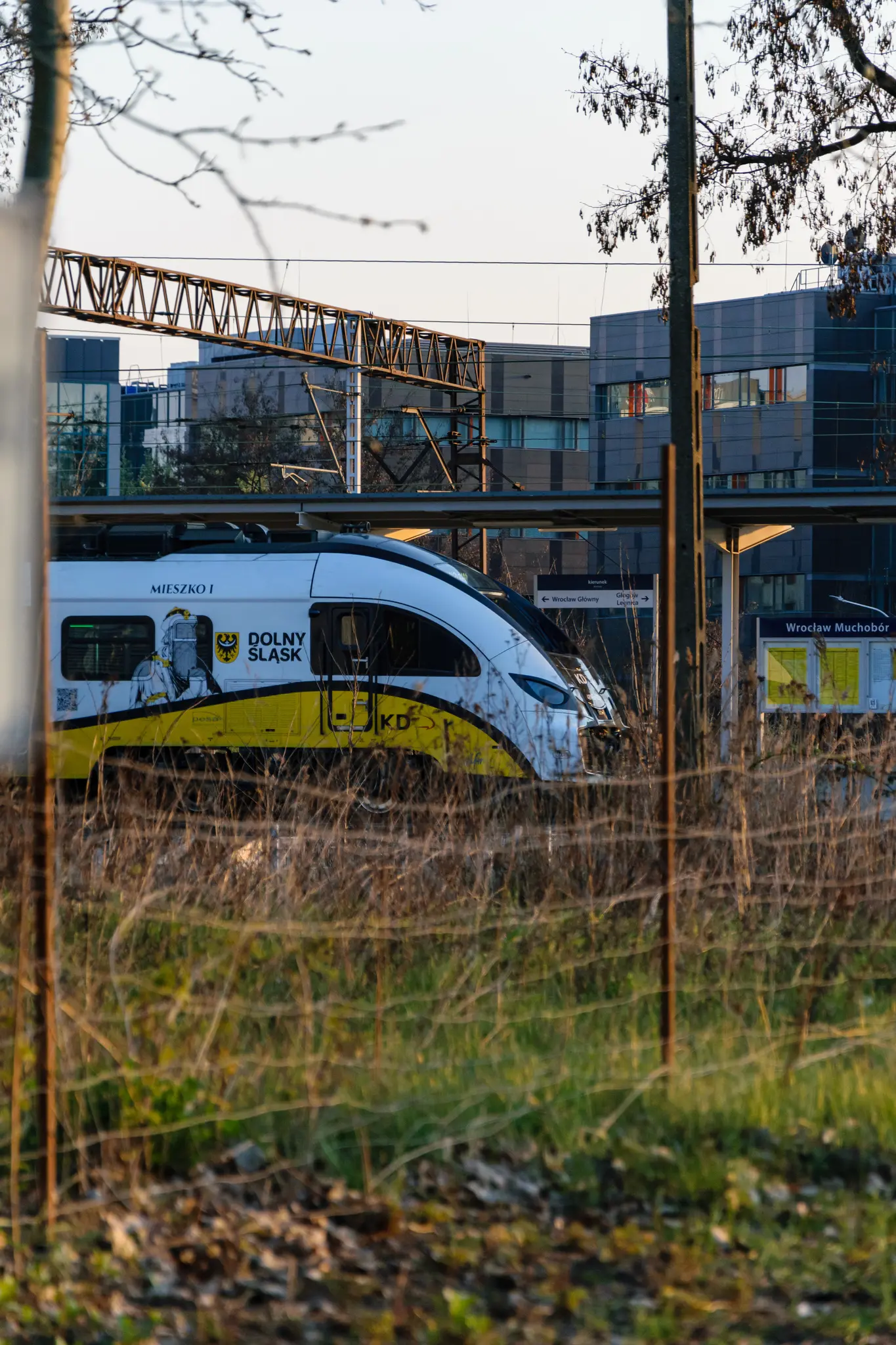 Yellow-white train stopped at a station, viewed from behind a barbed wire fence???