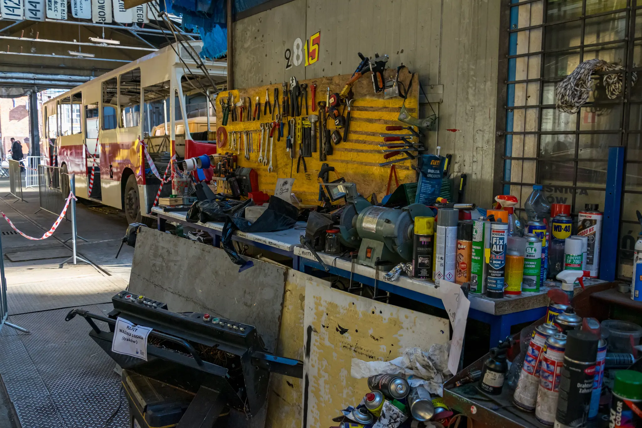 a workshop with various tools hanging on a yellow wall, a workbench with spray cans and various powertools, and a bus in the background