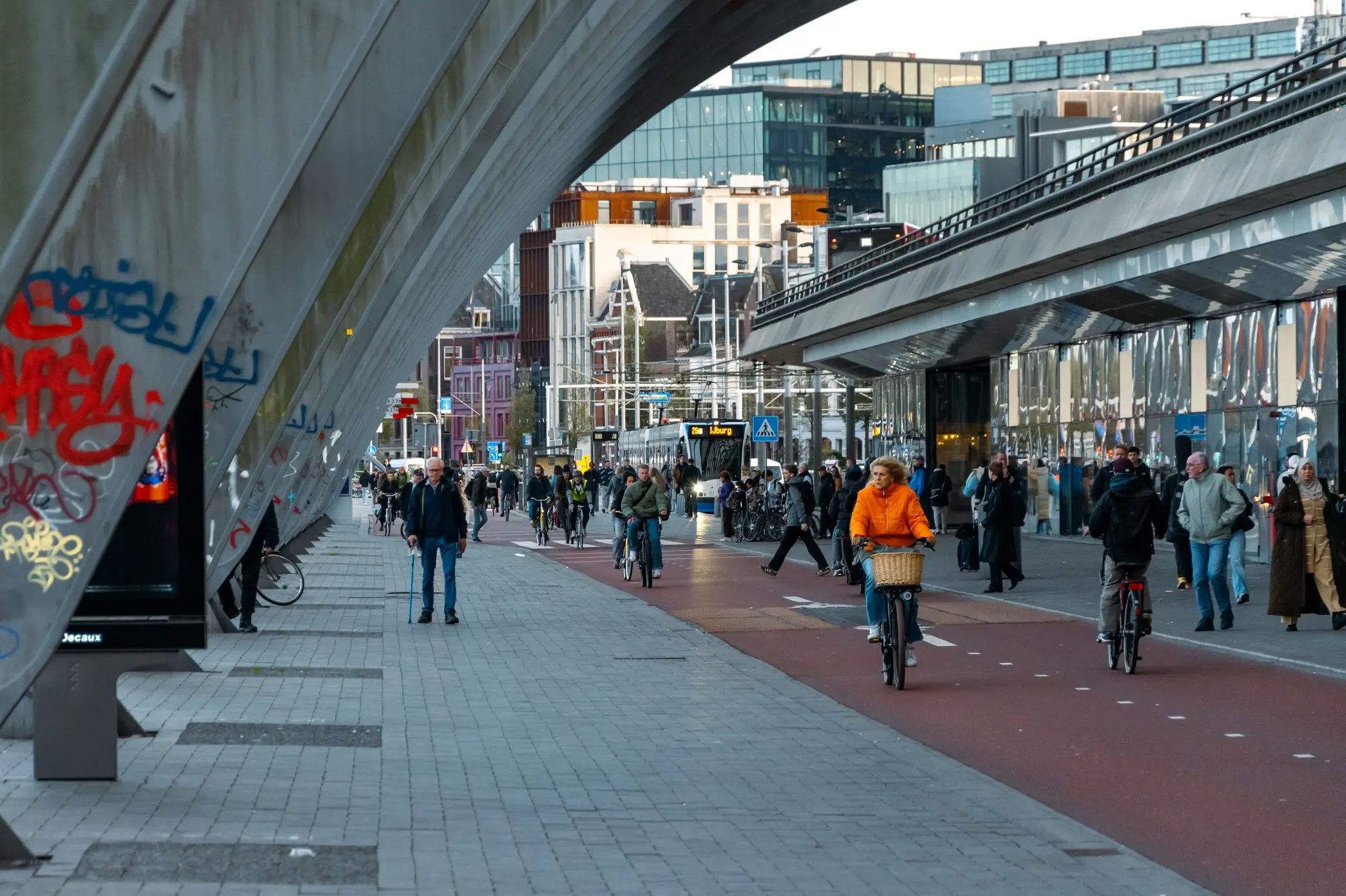Crowded path with a bike path embedded. There’s lots of people walking and biking, with tram waiting under modern arches of railway station