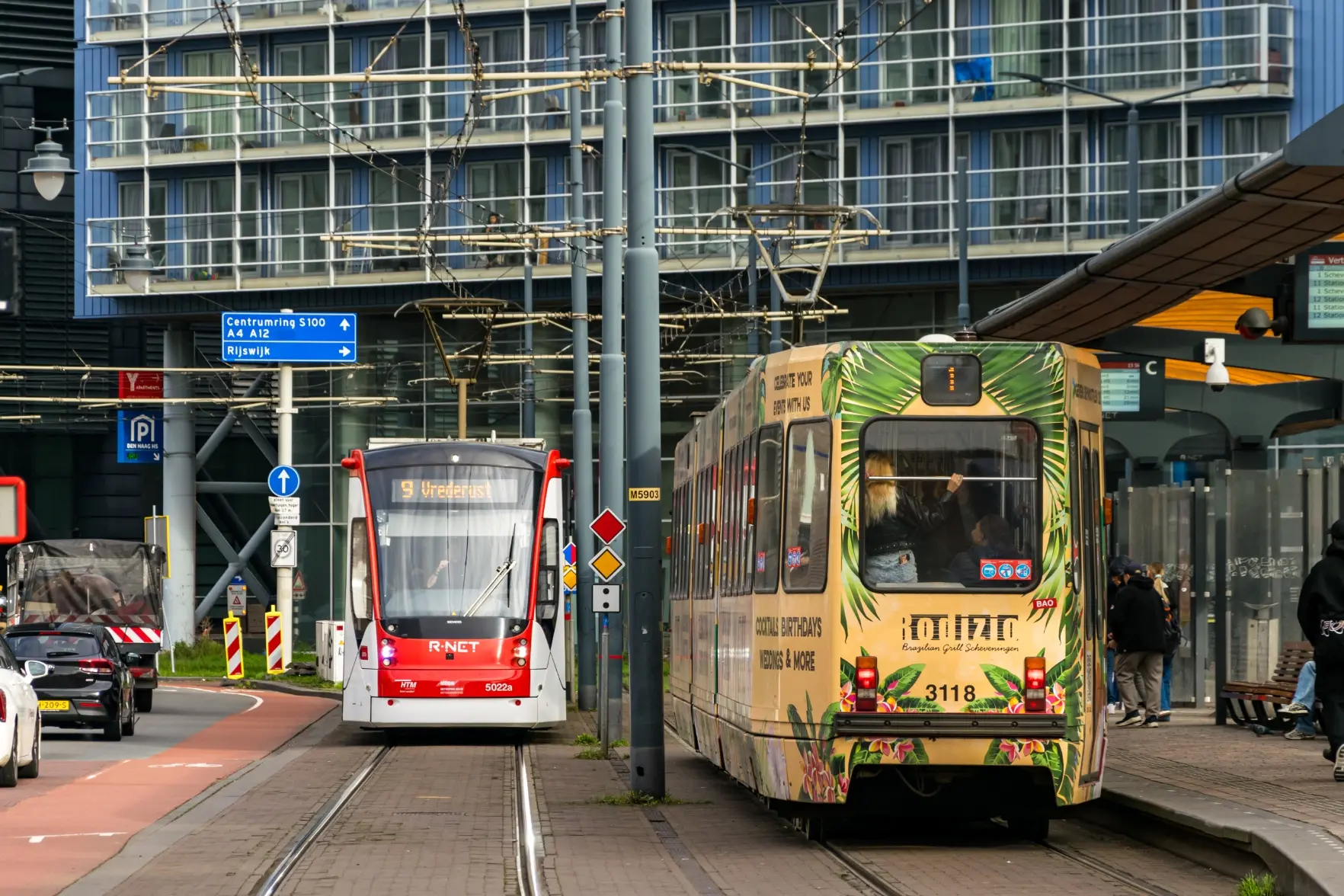 old tram in an ad livery at a platform, while other red modern tram is arriving soon