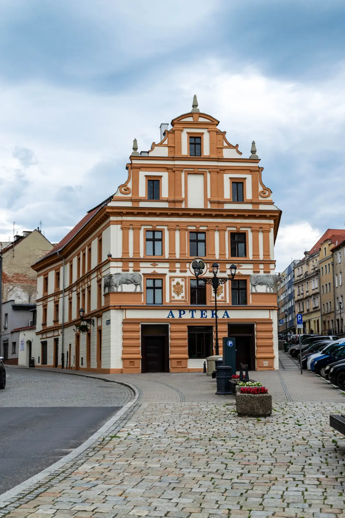 creamy building with lots of dark orange accents mostly around windows, has bulls on sides, there's text "Apteka"