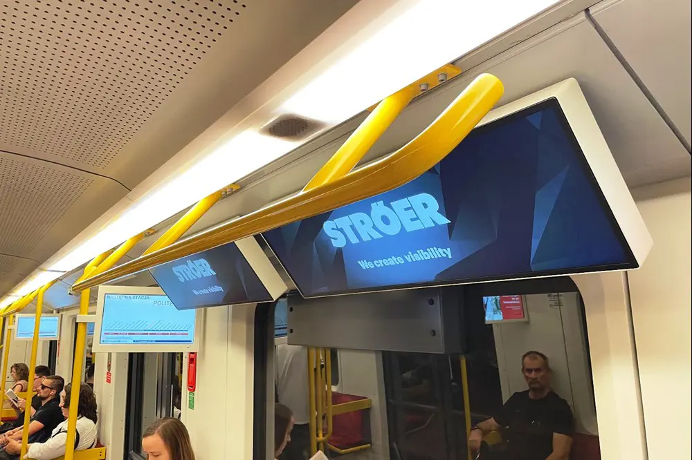 Grube ekrany reklamowe w pociągu metra, skierowane w pasażerów siedzących na wprost nich, oraz zasłaniają ekrany informacji pasażerskiej